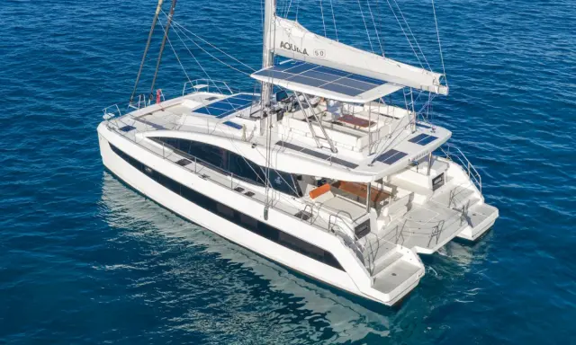 Un catamaran blanc, une véritable bicoque bateau, navigue sur une mer bleue. Ses voiles sont partiellement déployées, et des panneaux solaires brillent sur son toit.