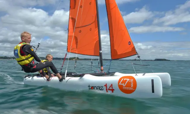 Deux jeunes garçons naviguent sur un petit voilier 2 places, leurs voiles oranges gonflées par le vent sous un ciel bleu et nuageux.