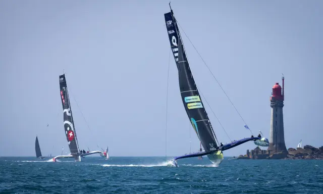 Le trimaran Koesio fend les vagues, une course intense près d'un phare.