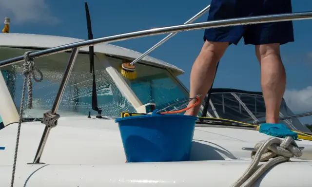 Un homme effectue l'entretien de bateau, lavant la proue avec une brosse et un seau bleu.