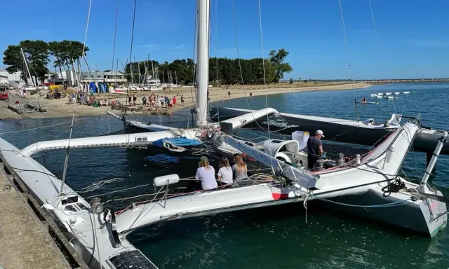 Le trimaran Flo, un bateau à trois coques, est amarré à quai. Des personnes sont à bord et sur la plage en arrière-plan.