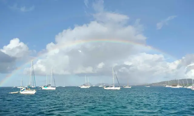Un arc-en-ciel sur une mer bleue, parsemée de voiliers. L'appel de l'aventure : tout quitter pour vivre sur un bateau.