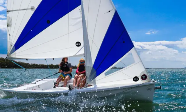 Deux jeunes femmes naviguent sur un voilier RS Venture par une journée ensoleillée. L'eau scintille sous le ciel bleu.