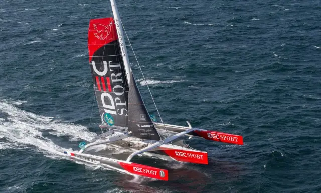 Le trimaran IDEC SPORT fend les vagues, une voile rouge et noire ornée d'un oiseau blanc.