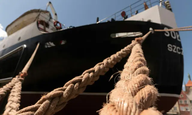 Gros plan sur des cordages épais assurant l'amarrage d'un bateau noir et blanc, avec des bâtiments colorés en arrière-plan.