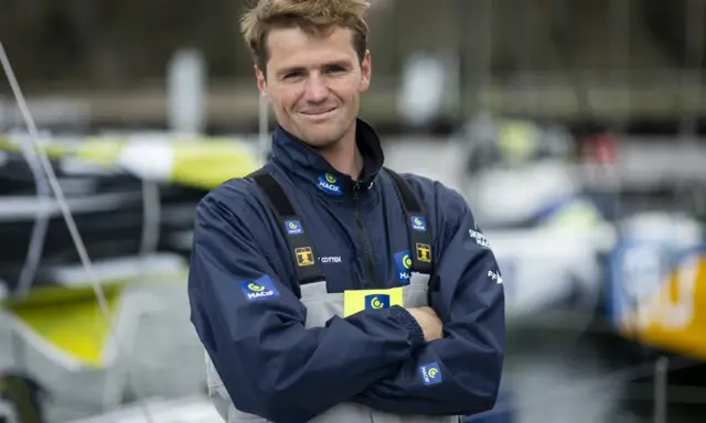 L'athlète marin Lois Berrehar, souriant, porte une veste bleue MACIF avec des logos. Derrière lui, des voiliers colorés.