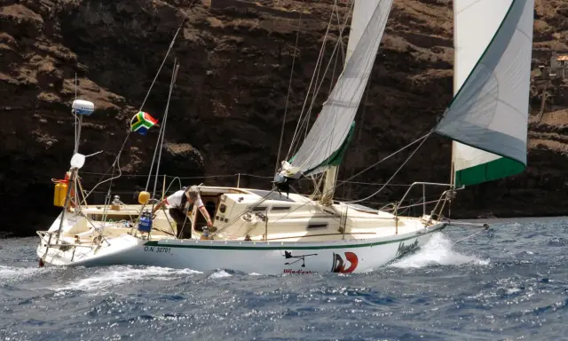 Un voilier cap horn, le "Windicator", navigue près d'une côte rocheuse. Le drapeau sud-africain flotte fièrement.