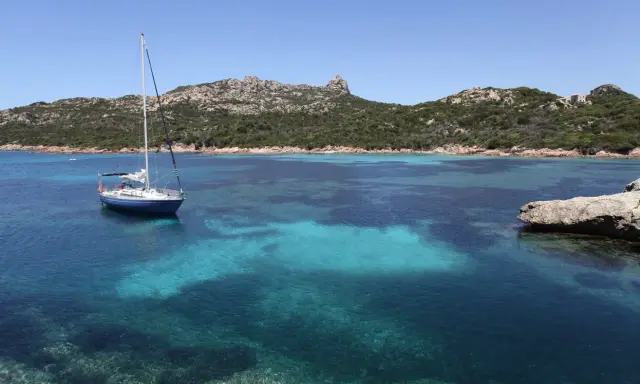 Un voilier bleu ancré dans les eaux turquoise de Cap Corse, avec des collines verdoyantes en arrière-plan sous un ciel clair.