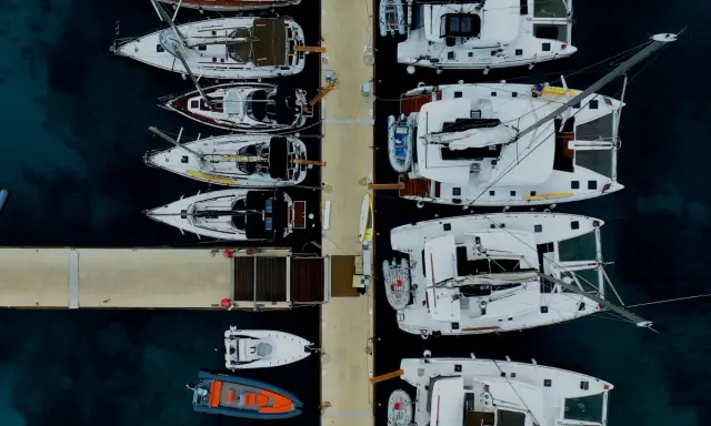 Vue aérienne d'un port avec plusieurs yachts à voile blancs amarrés, certains catamarans, et des annexes gonflables.