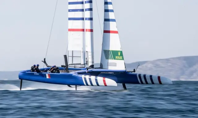 Catamaran bleu et blanc, voiles rayées, en pleine action. La **vitesse catamaran** est impressionnante, les foils hors de l'eau.