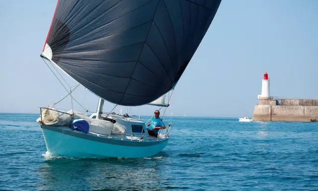 Un voilier bleu avec une grande voile noire navigue près d'un phare. Le skipper, portant un t-shirt bleu, semble observer un ovni 25 dans le ciel clair.