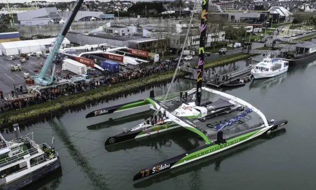 Le trimaran Sodebo Ultim 3, prêt pour la course, est amarré dans un port, entouré de spectateurs et de grues.