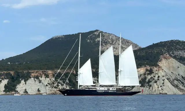 Le yacht de Bezos, un voilier majestueux, navigue en Méditerranée, ses voiles blanches déployées devant des falaises escarpées.