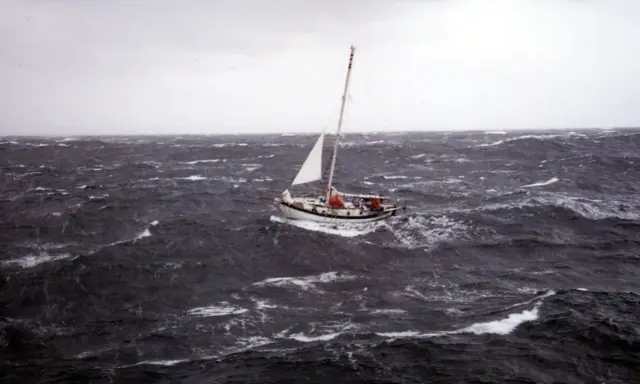 Un voilier blanc lutte contre les vagues déchaînées dans une mer agitée. Le ciel est gris et menaçant, annonçant une tempête.