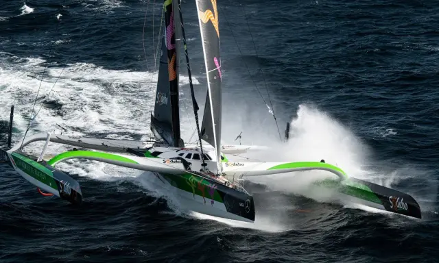Le trimaran Sodebo, un bateau ultime, fend les vagues sombres, projetant des gerbes d'eau dans une course effrénée.