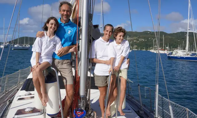 Famille souriante sur un voilier, un moment clé de leur récit de voyage en voilier. La mer bleue et les voiliers en arrière-plan complètent ce tableau idyllique.
