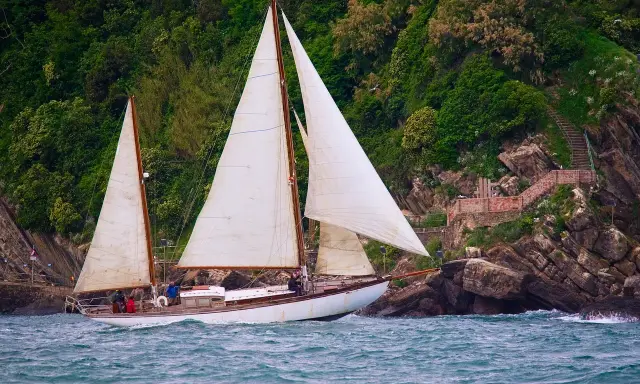 Un magnifique voilier 2 mâts navigue près d'une côte rocheuse verdoyante.