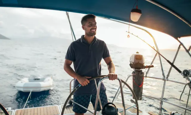 Un homme souriant barre un bateau, profitant du soleil couchant. Un petit bateau gonflable est attaché à l'arrière.