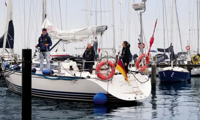 Trois personnes sur un bateau, prêtes à appareiller. Le drapeau allemand flotte fièrement. L'erre bateau est calme.