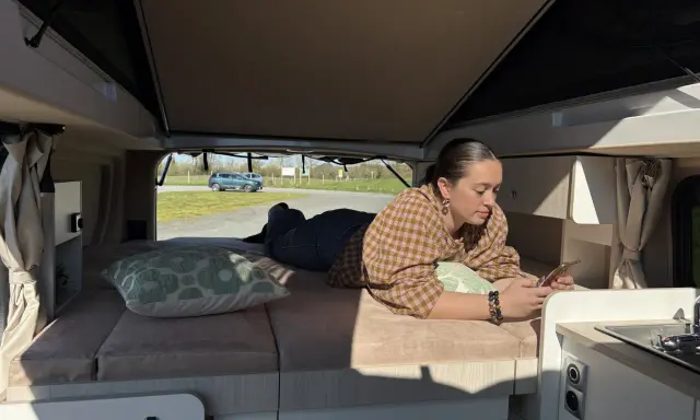 Une jeune femme se détend sur le lit de son camping-car j80 interieur, regardant son téléphone. Dehors, le soleil brille.