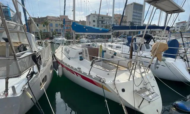 Un voilier First 32, amarré dans un port, avec d'autres bateaux et des bâtiments en arrière-plan.