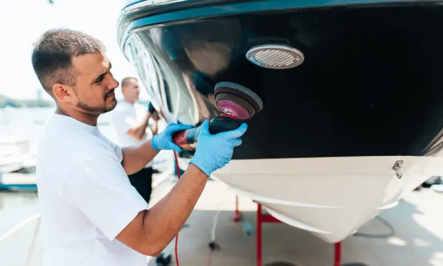 Un homme polit la coque d'un bateau. Il utilise une polisseuse pour un travail de précision, peut-être en prévision d'une révision moteur inboard.
