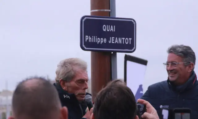 Un panneau "Quai Philippe JEANTOT" honore le navigateur. Philippe JEANTOT, entouré, parle au micro.