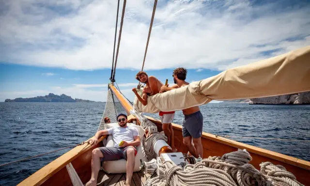 Trois amis profitent du soleil sur un voilier, rêvant d'un tour du monde. L'eau bleue et les falaises en arrière-plan ajoutent à l'aventure.
