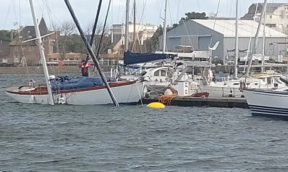 Un voilier blanc et bois, bâché, est partiellement immergé dans un port. Un homme en gilet rouge est à bord.