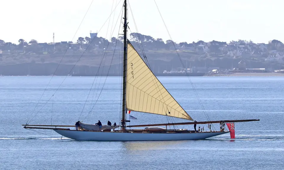 Un élégant voilier, surnommé "mariquita", navigue sur une mer bleue, sa grande voile beige déployée. Une côte verdoyante bordée de maisons se dessine à l'horizon.