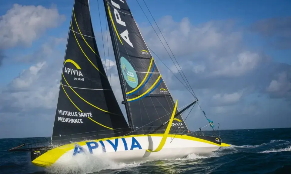 Le voilier Apivia, skippé par Alex Thomson, fend les vagues sous un ciel bleu et nuageux.