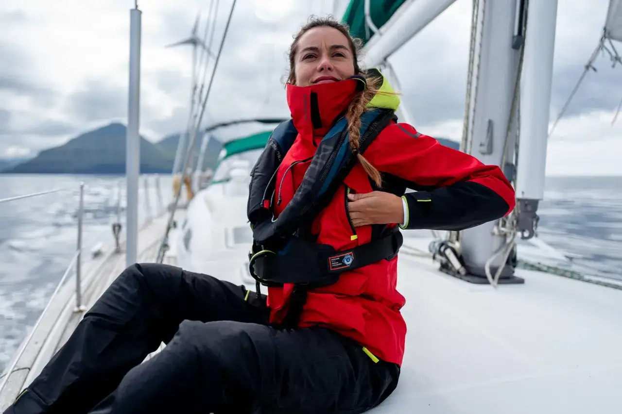 Une femme en gilet de sauvetage rouge et noir sur un voilier. Elle ajuste sa veste, réfléchissant à quel gilet de sauvetage choisir pour naviguer en toute sécurité.