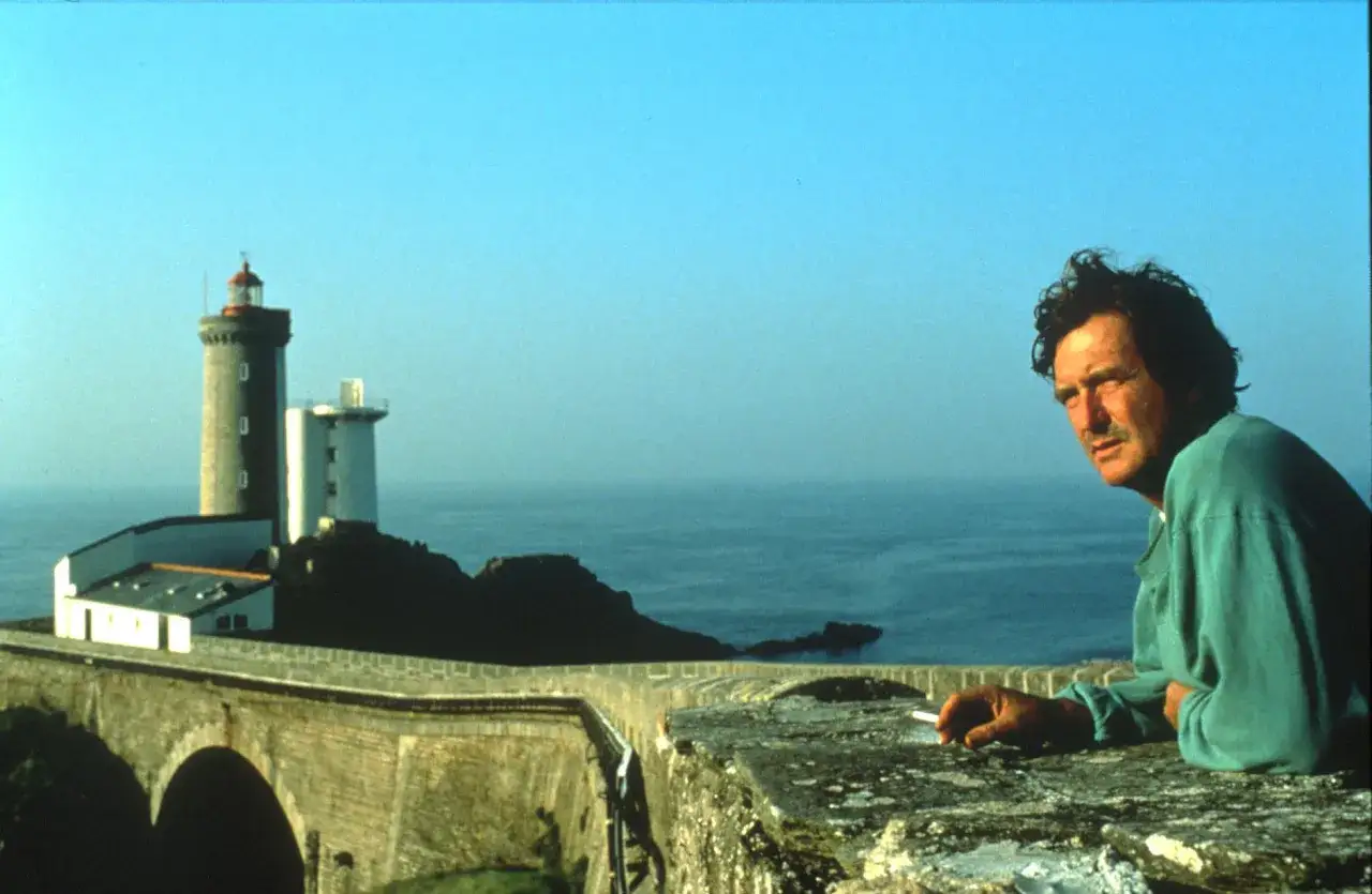Olivier de Kersauson jeune, pensif, fume une cigarette près d'un phare sur une falaise bretonne.