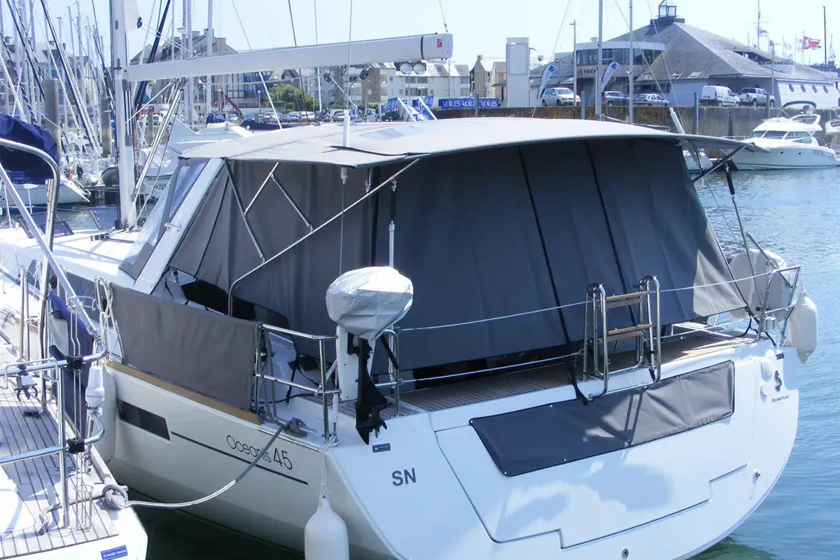 Le cockpit d'un bateau de plaisance, l'Océanis 45, est protégé par une bâche grise. Des mâts et d'autres bateaux sont visibles en arrière-plan.