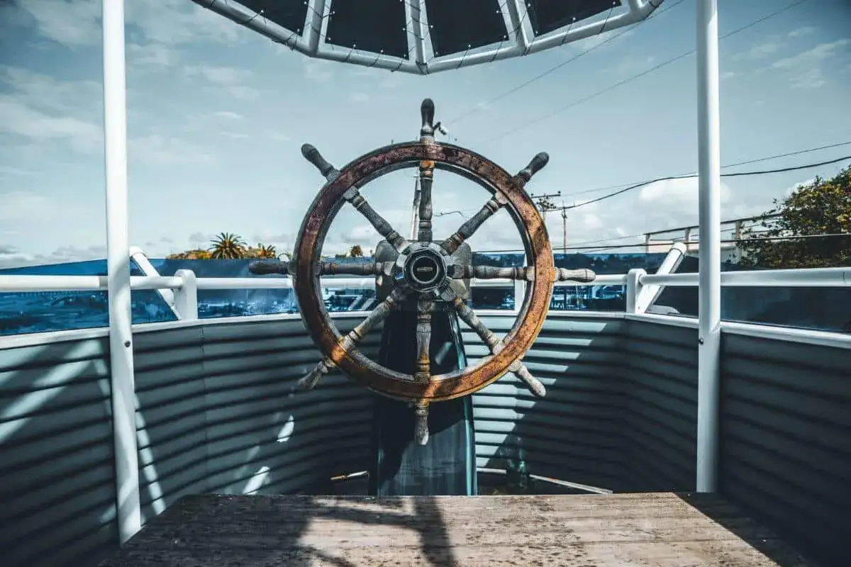 Une barre à roue en bois, usée par le temps, trône sur un pont. Le ciel bleu et les nuages annoncent une belle journée en mer.