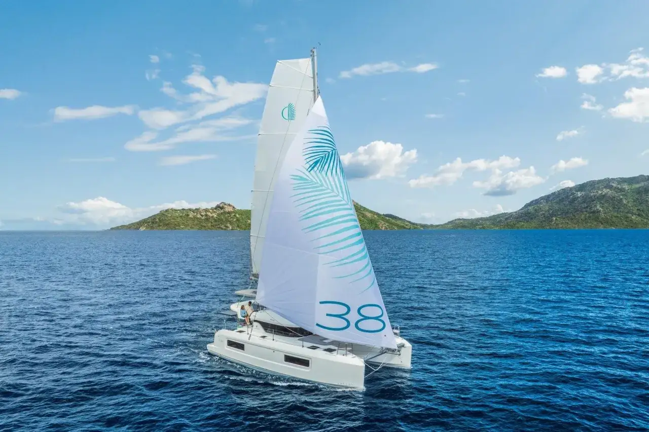 Un catamaran blanc navigue sur une mer bleue, voiles gonflées. Sébastien Roubinet admire le paysage côtier verdoyant.