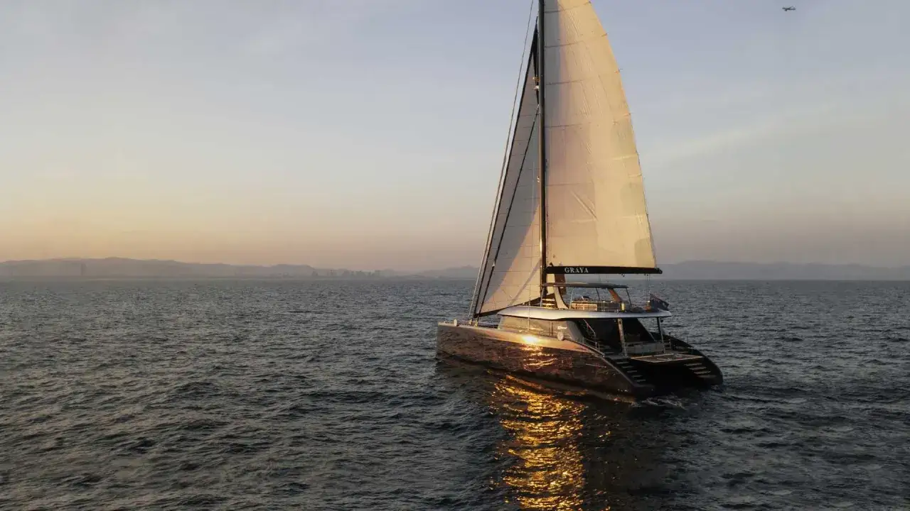 Un luxueux prao bateau navigue sur une mer calme au coucher du soleil, ses voiles illuminées par la lumière dorée.