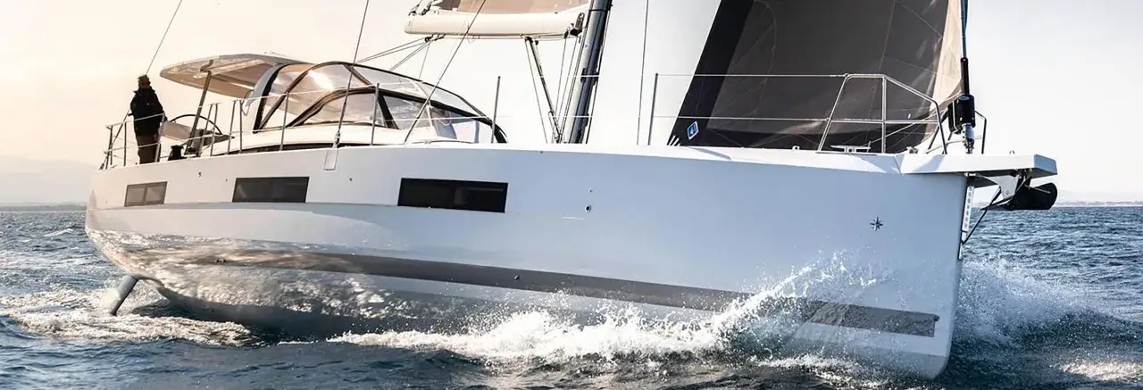 Un voilier transportable hauturier fend les vagues, son étrave blanche projetant des gerbes d'eau sous un ciel clair.