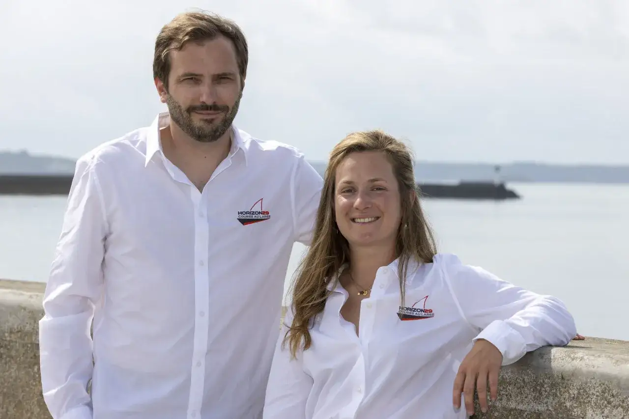 Élodie Bonafous et un homme souriants, portant des chemises blanches avec un logo de voile. Ils posent près de l'eau.