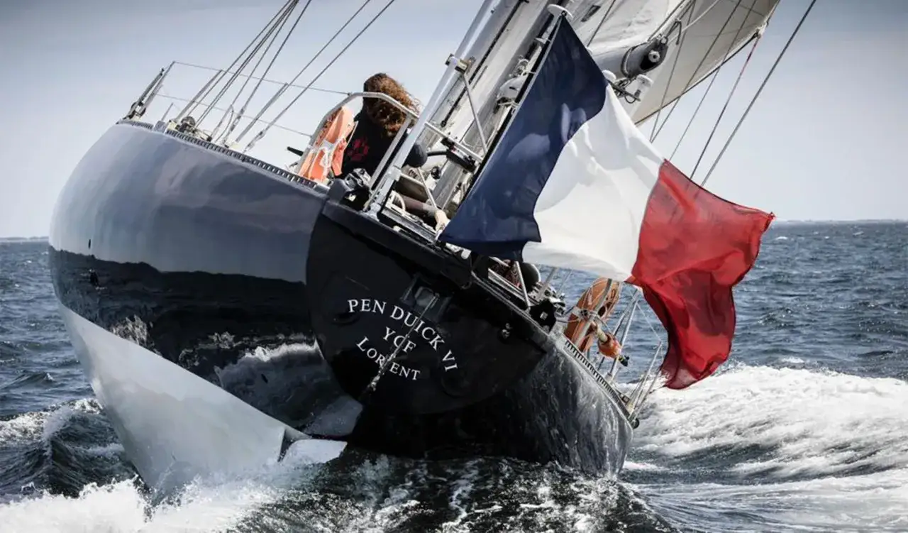Le voilier Pen Duick VI, avec le drapeau français, navigue sur une mer agitée. Marie Tabarly, à la barre, semble apprécier la puissance du bateau.