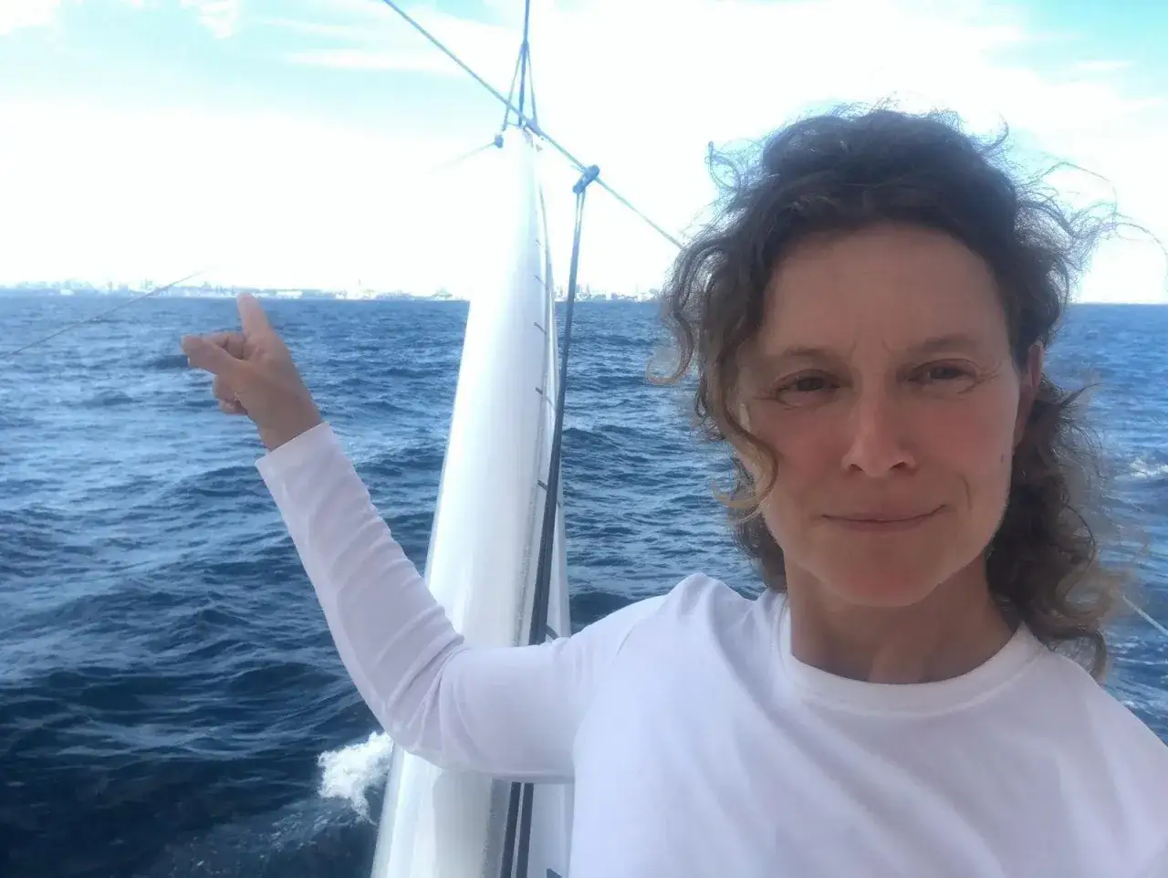 Isabelle Joschke, souriante, pointe le doigt vers l'horizon depuis un voilier, la mer bleue et le ciel clair en arrière-plan.