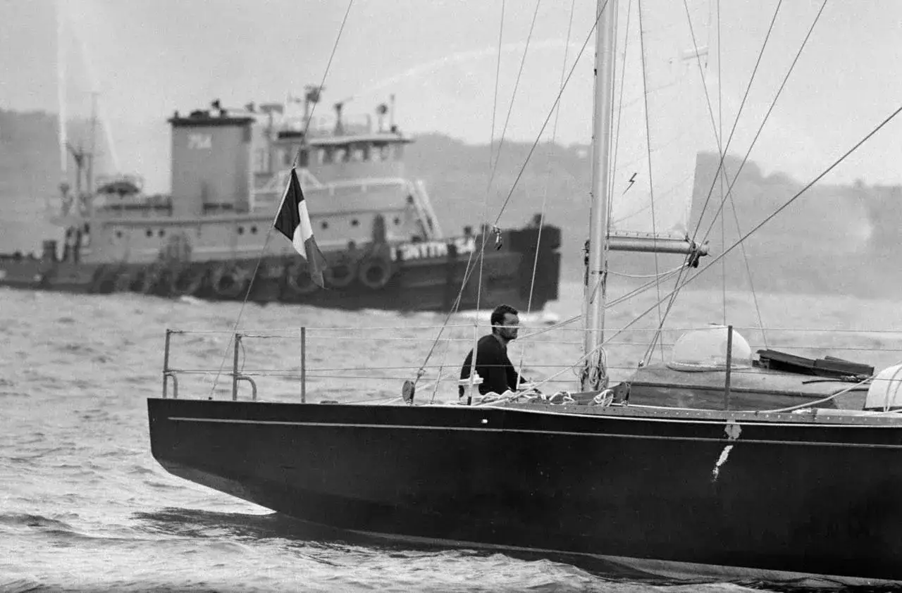 Jacques Arthaud, seul à la barre de son voilier, navigue avec un remorqueur en arrière-plan. Le drapeau français flotte fièrement.