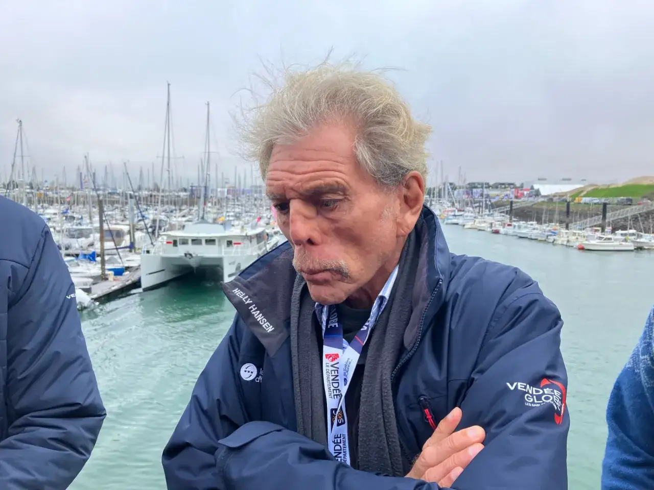 Philippe Jeantot, le regard pensif, devant un port rempli de voiliers. Il porte une veste Helly Hansen, rappelant peut-être un ancien accident de voile.