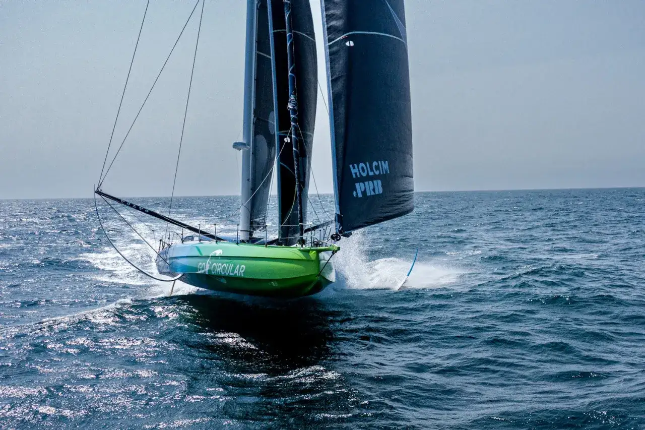 Un bateau de course, tel un tapis volant, fend les vagues bleues. Ses voiles noires portent