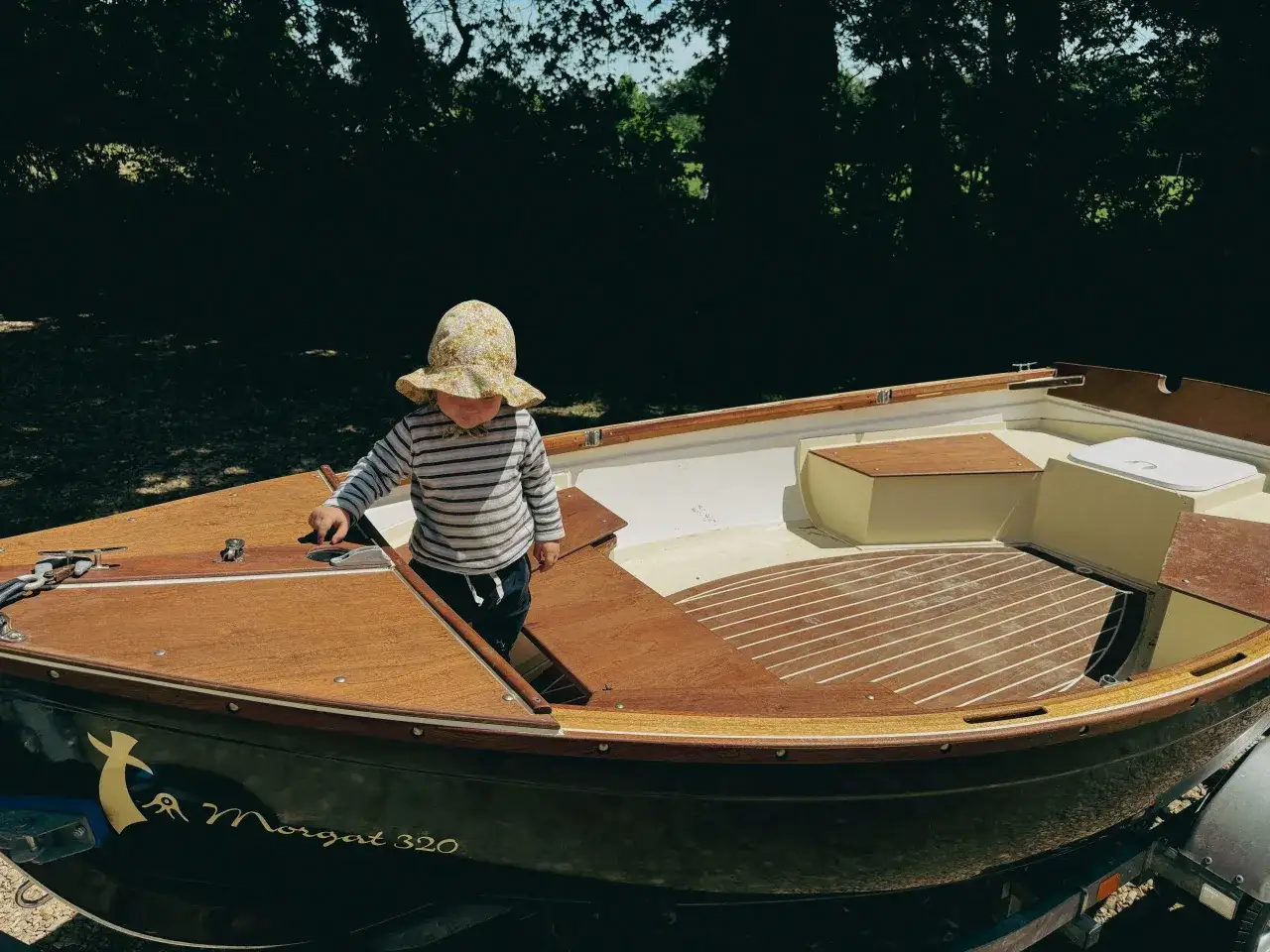 Un enfant en chapeau fleuri explore l'intérieur d'un bateau en bois, le Morgat 320, prêt pour l'aventure.