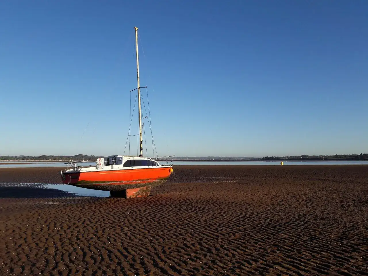 Un voilier Biloup Club orange échoué sur une plage de sable fin, sous un ciel bleu clair.