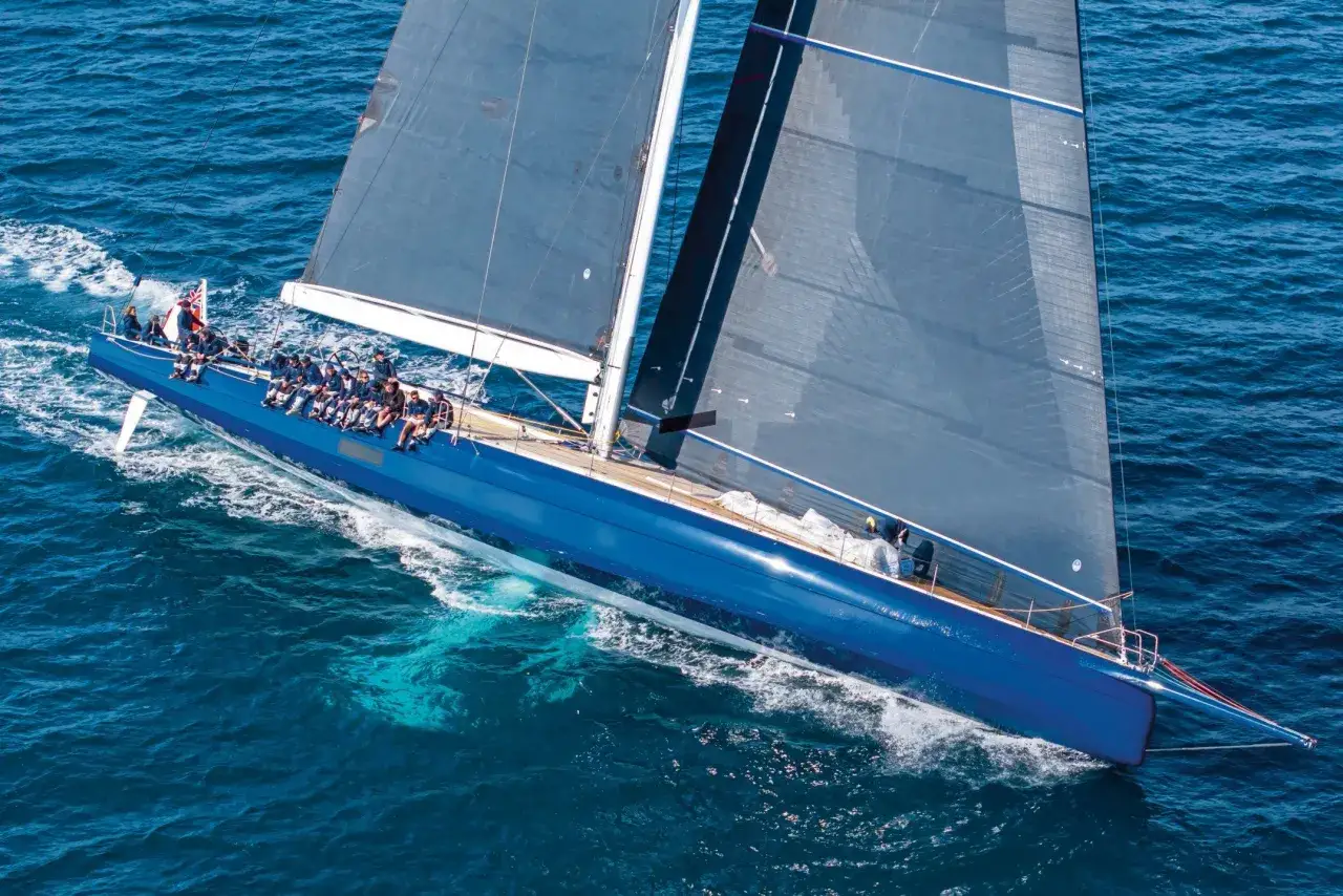 Un voilier bleu vif glisse sur l'eau, ses voiles sombres déployées comme un tapis volant 3. L'équipage est visible sur le pont.