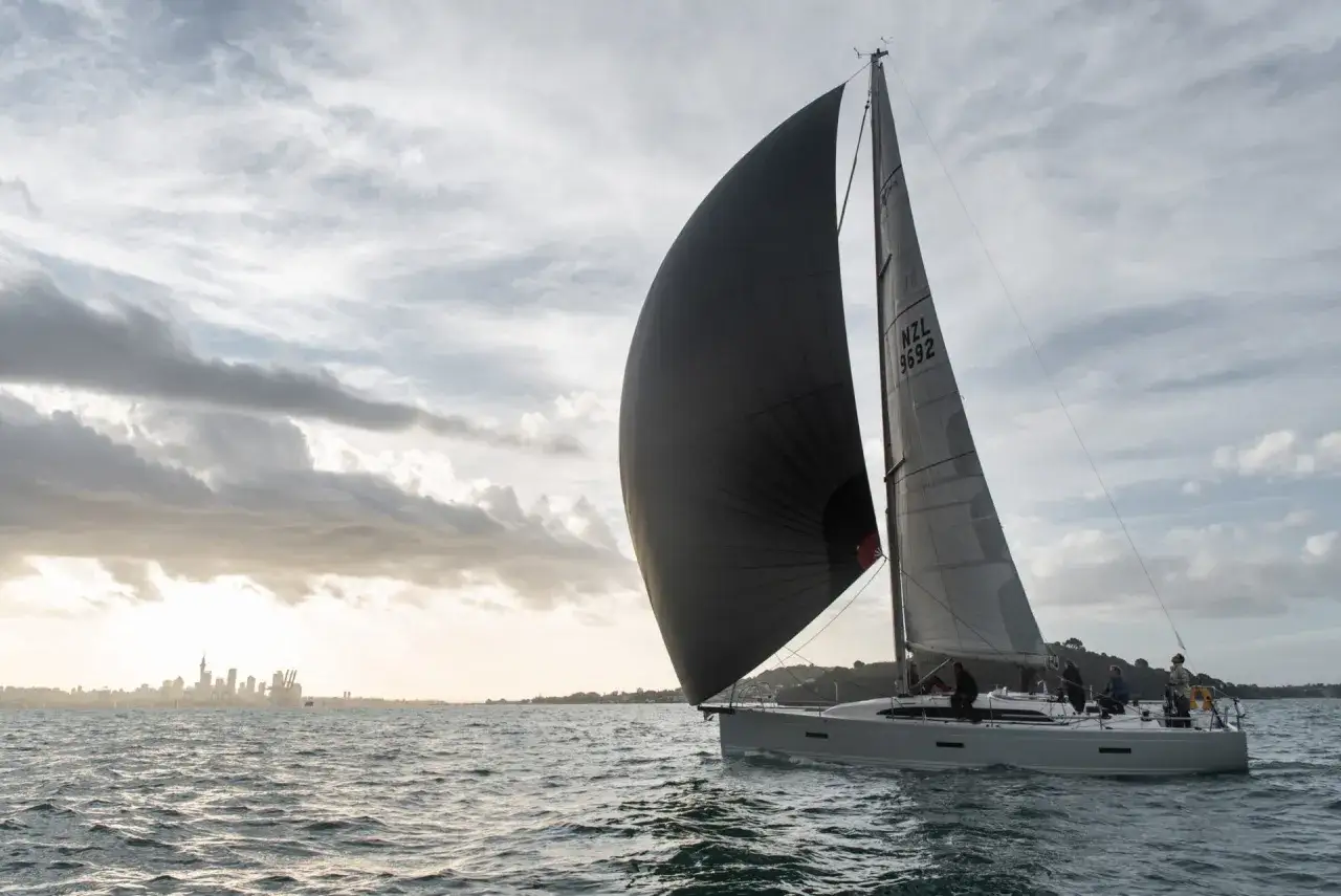 Un voilier navigue avec un grand gennaker noir gonflé par le vent, contrastant avec le spinnaker gris. La ville d'Auckland se dessine à l'horizon.
