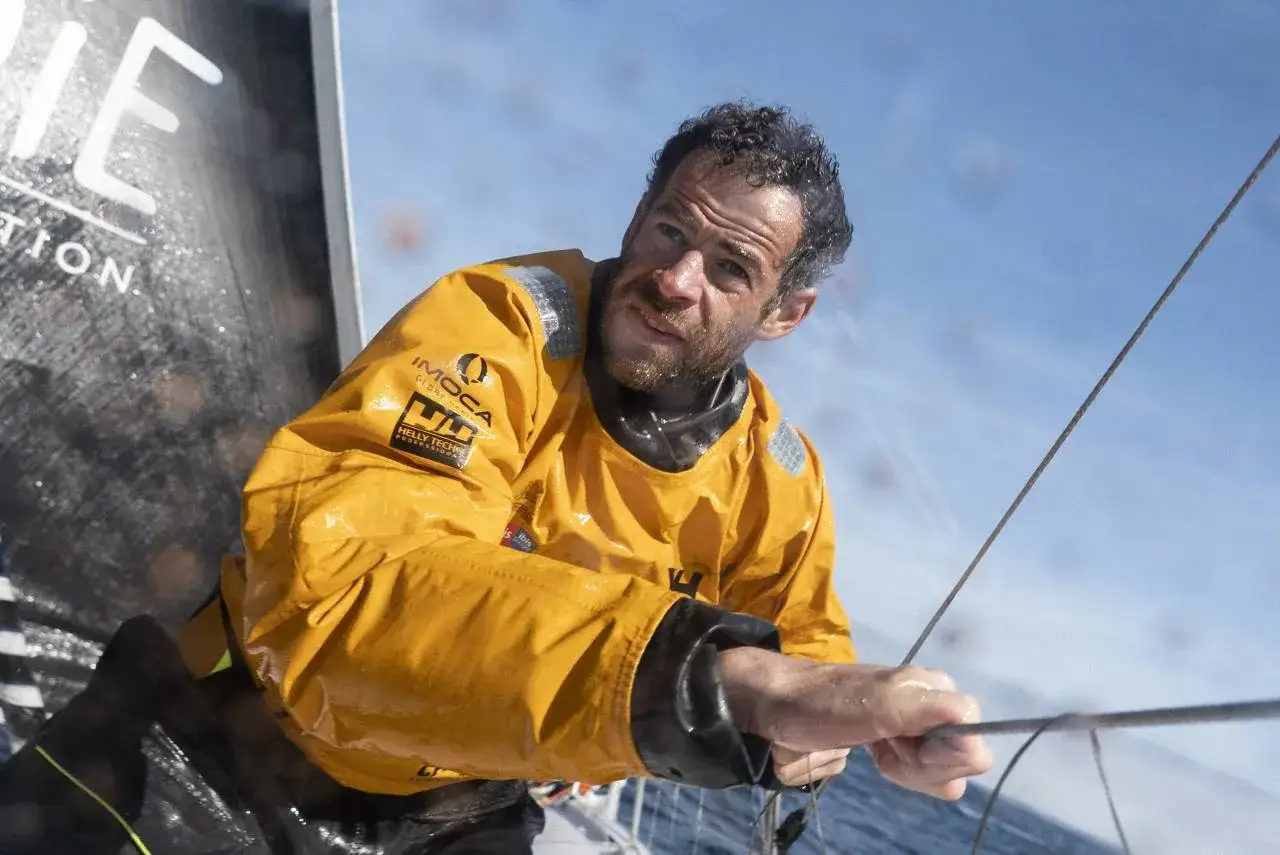 Nicolas d'Estais, marin concentré, manœuvre une voile sous un ciel bleu, vêtu d'une veste jaune imperméable.