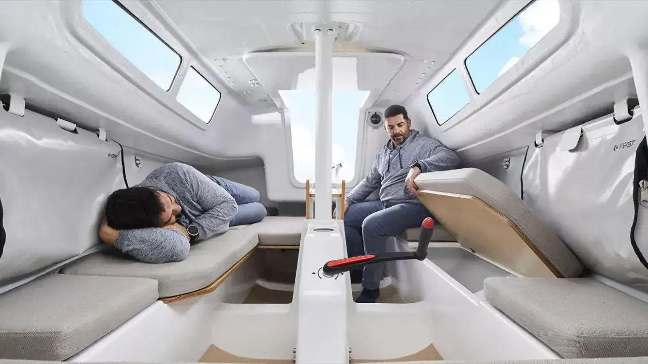 Intérieur du FIRST 24 : un homme dort sur une banquette, un autre est assis, prêt à manœuvrer. Lumière naturelle filtrant par les hublots.
